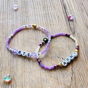 May include: Two beaded bracelets with the words "BTS" and "ARMY" spelled out in white letter beads. The bracelets are purple and white with gold and dark red accents. The bracelets are on a wooden surface.