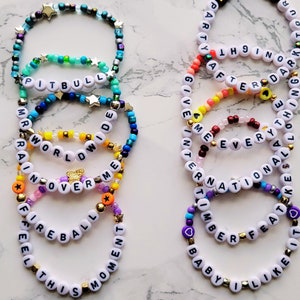 May include: A set of ten colorful beaded bracelets with different words spelled out on them. The words include "Pitbull", "Mr Worldwide", "Rain Over Me", "Feel This Moment", "Party All The 9 In The Dark", "Give Me Every Thing", "International Feat", "Baby I Like It", and more.