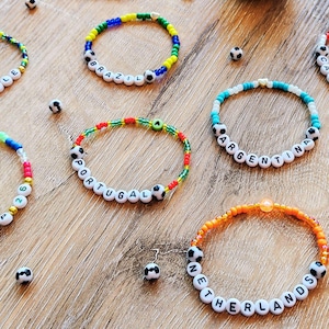 Könnte beinhalten: Eine Sammlung von Armbändern mit Perlen, die Ländernamen und Fussballakzente tragen. Die Armbänder sind in verschiedenen Farben gehalten und werden auf einer Holzoberfläche präsentiert.
