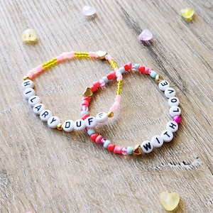 May include: Two beaded bracelets with white letter beads spelling "HILARY DUFF" and "LOVE WITH". The bracelets feature gold heart charms and are accented with pink, red, and yellow beads. Scattered heart-shaped candies are also visible.
