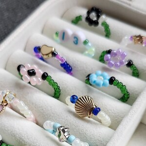 以下が含まれることがあります： ハート、花、星、貝殻、数字の「13」など、さまざまなチャームが付いたカラフルなビーズリングのコレクション。リングは、右下に「Blank Space Bracelets」というテキストが表示されている白いリングホルダーに展示されています。