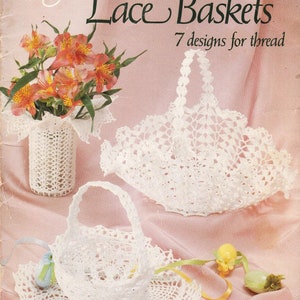 Op de afbeelding: Een vintage handwerkpatroonboek voor gehaakte kantmanden. De omslag heeft een roze achtergrond met witte kantmanden en de titel "Crocheted Lace Baskets". De tekst "7 designs for thread" is ook inbegrepen.