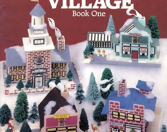 May include: Plastic canvas pattern for a Christmas village, featuring a red brick building with a white roof, a green building with a white roof and a red door, a pink building with a white roof and a red door, and a blue building with a white roof and a red door. The pattern is titled "Home for Christmas Village" and is by Don FranzMeier.