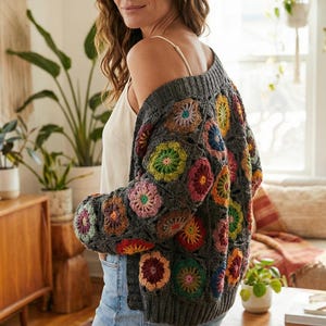 Puede incluir: Un cárdigan de ganchillo gris con coloridos estampados florales. El suéter abierto presenta una variedad de diseños florales en tonos de rosa, amarillo, verde y naranja. El cárdigan se lleva sobre una camiseta sin mangas blanca y vaqueros azules.