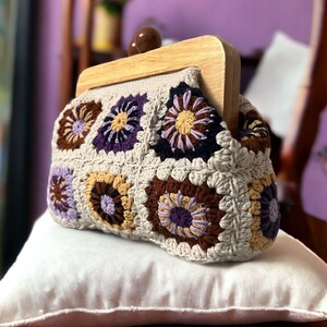 Può includere: Una pochette a uncinetto con una cornice in legno. La borsa è realizzata in filato beige con accenti marroni, viola e gialli. La borsa ha un motivo floreale.
