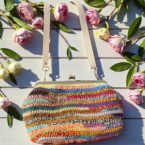 May include: A colorful, crocheted handbag with a metal clasp and cream-colored straps. The bag features a vibrant mix of colors, including pink, yellow, blue, and orange. The background includes pink roses and green leaves on a white wooden surface.