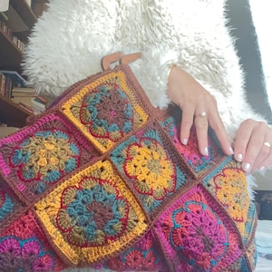 Può includere: Una borsa a mano in crochet con manico in pelle marrone. La borsa è realizzata con quadrati colorati con un motivo floreale in tonalità marrone, rosa, giallo e turchese.