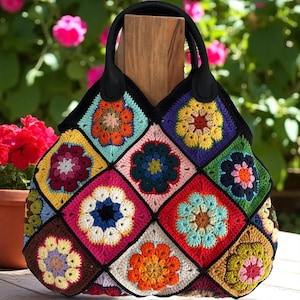 May include: A colorful, handmade crochet tote bag. The bag features a patchwork design of floral squares in various colors, including red, yellow, blue, and green. It has a black handle and a black border around the squares. The bag is sitting on a wooden surface.