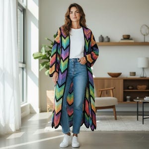 Op de afbeelding: Een lange, open gehaakte vest met een levendig chevronpatroon. Het kleurrijke ontwerp heeft een spectrum aan regenboogtinten tegen een zwarte achtergrond. Het vest is knielang en heeft lange mouwen.