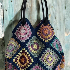 Può includere: Una borsa a mano all'uncinetto con sfondo blu scuro e quadrati di granny colorati. La borsa ha manici in pelle nera.