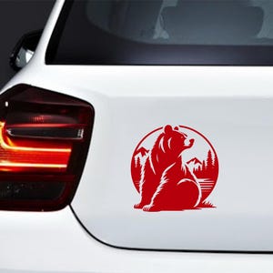 May include: Red bear silhouette sticker on a white car. The sticker features a bear sitting in front of a mountain and forest scene, enclosed in a circular frame. The design is a simple, bold graphic.