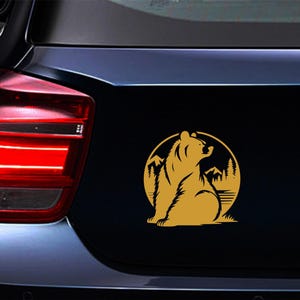 May include: A gold-colored bear silhouette sticker on a black car surface. The bear is seated, looking upwards, within a circular frame featuring mountains and trees. The sticker is affixed to the vehicle's rear.