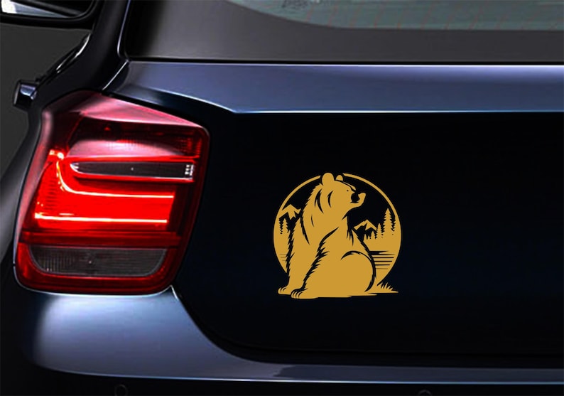 May include: A gold-colored bear silhouette sticker on a black car surface. The bear is seated, looking upwards, within a circular frame featuring mountains and trees. The sticker is affixed to the vehicle's rear.