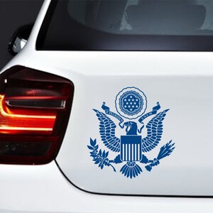 Pode incluir: Um adesivo azul e branco do Grande Selo dos Estados Unidos da Am&eacute;rica em um carro branco. O selo apresenta uma &aacute;guia com as asas abertas, um escudo com treze listras e as palavras "E Pluribus Unum".