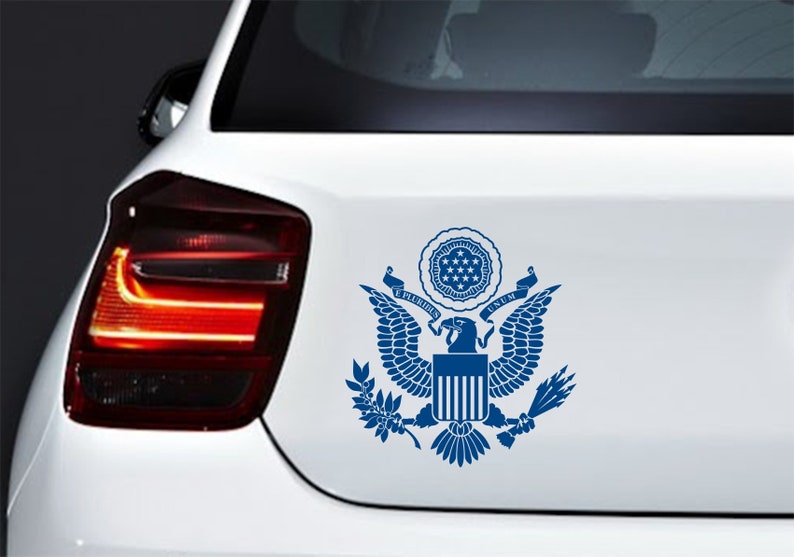Pode incluir: Um adesivo azul e branco do Grande Selo dos Estados Unidos da Am&eacute;rica em um carro branco. O selo apresenta uma &aacute;guia com as asas abertas, um escudo com treze listras e as palavras "E Pluribus Unum".