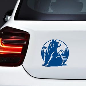 May include: A blue vinyl decal of a bear sitting in front of a mountain and forest scene, inside a circular frame. The decal is affixed to the white rear of a car.