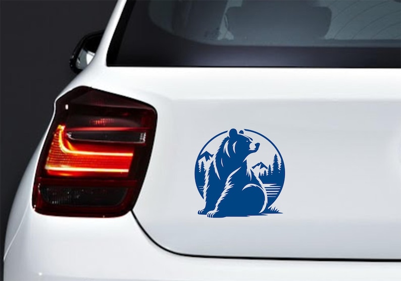 May include: A blue vinyl decal of a bear sitting in front of a mountain and forest scene, inside a circular frame. The decal is affixed to the white rear of a car.