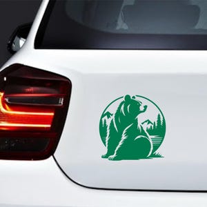May include: Green bear sticker on a white car. The sticker features a bear silhouette within a circular frame, with mountains and trees in the background. The bear is sitting and looking to the right. The sticker is applied to the car's rear.