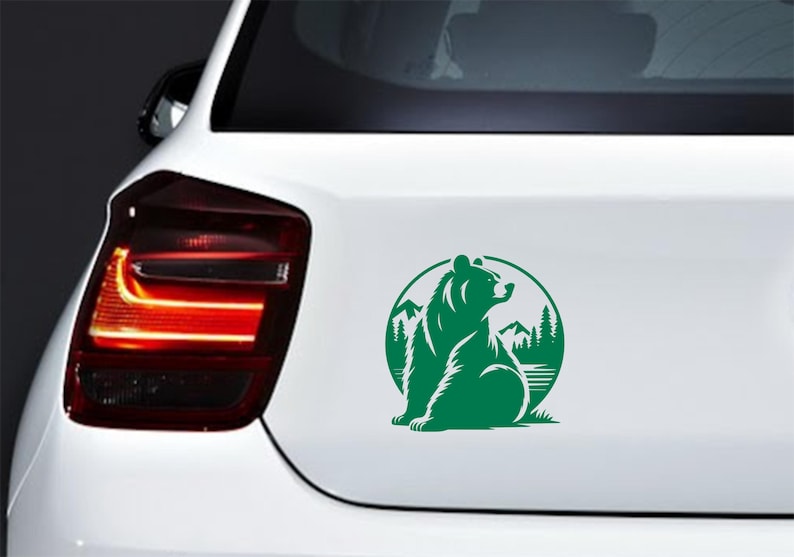May include: Green bear sticker on a white car. The sticker features a bear silhouette within a circular frame, with mountains and trees in the background. The bear is sitting and looking to the right. The sticker is applied to the car's rear.
