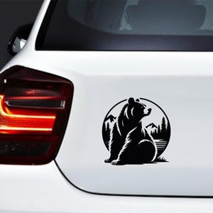 May include: Black and white vinyl decal of a bear sitting in front of a mountain and forest scene, inside a circle. The decal is affixed to the rear of a white vehicle.