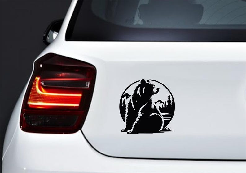 May include: Black and white vinyl decal of a bear sitting in front of a mountain and forest scene, inside a circle. The decal is affixed to the rear of a white vehicle.