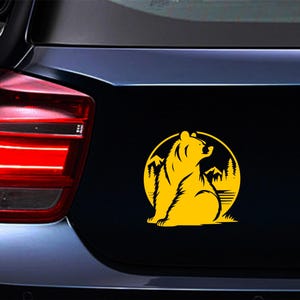 May include: A yellow bear silhouette sticker on a black surface. The bear is seated inside a yellow circle with mountains and trees. The sticker is likely for a car or vehicle.