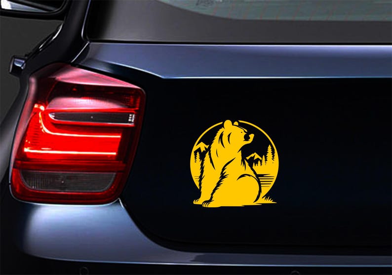 May include: A yellow bear silhouette sticker on a black surface. The bear is seated inside a yellow circle with mountains and trees. The sticker is likely for a car or vehicle.