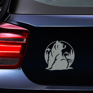 May include: A gray bear sticker on a car's trunk. The bear is silhouetted, seated, and looking upwards within a circular frame. The frame includes mountain and tree graphics, suggesting a nature theme.