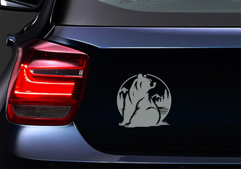 May include: A gray bear sticker on a car's trunk. The bear is silhouetted, seated, and looking upwards within a circular frame. The frame includes mountain and tree graphics, suggesting a nature theme.