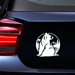 May include: White bear silhouette sticker on a black car surface. The bear is inside a circle with mountain and tree details. The sticker is likely for a car or vehicle.