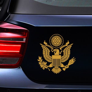 Pode incluir: Um adesivo dourado do Grande Selo dos Estados Unidos da Am&eacute;rica em um carro azul. O selo apresenta uma &aacute;guia com as asas abertas, um escudo com listras verticais e o lema "E Pluribus Unum".