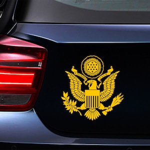 Pode incluir: Um adesivo dourado do Grande Selo dos Estados Unidos, com uma &aacute;guia de asas abertas, um escudo com listras verticais e uma faixa que diz "E PLURIBUS UNUM".