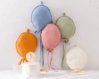 Peut inclure: Cinq décorations de ballons en tissu de différentes couleurs, dont bleu, rose, vert et orange, avec des ficelles qui pendent. Un gâteau blanc avec un numéro un en bois sur le dessus est au premier plan.