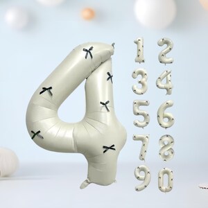 May include: A set of cream-coloured number balloons, including the number four, decorated with black bow accents. Additional numbers one through zero are also displayed. These balloons are suitable for birthday celebrations or other special events.