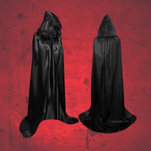 May include: Two black hooded capes made from a shiny, satin-like fabric. The capes have long, flowing designs, with one tied at the neck and the other with a simple hood. The background is a textured red surface.