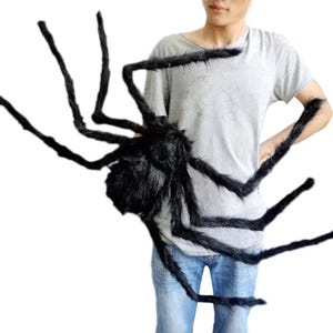 Op de afbeelding: Een grote, zwarte, harige spinnenprop met lange, gelede poten. De spin heeft een rond lichaam en is ontworpen voor Halloween of thema-decoraties. De spin wordt door een persoon vastgehouden.