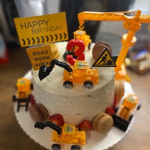 Könnte beinhalten: Ein runder Geburtstagskuchen, verziert mit Spielzeug-Baustellenfahrzeugen, Makronen und Erdbeeren. Der Kuchen hat ein gelbes Schild mit der Aufschrift "Happy Birthday" und "Road Work Ahead". Eine rote Kerze mit der Zahl 3 befindet sich oben.