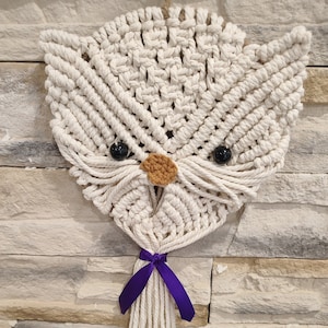 Può includere: Un arazzo da parete a forma di gufo in macramè. Il gufo è realizzato con cordoncino di cotone bianco sporco, con occhi di perline nere e un piccolo naso marrone. Un nastro viola è legato in basso, con il cordoncino che forma una nappa. Il gufo è montato su un muro di pietra strutturato.