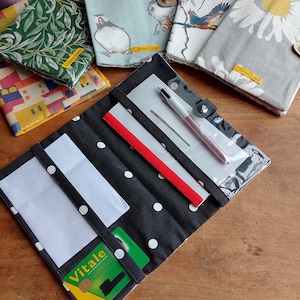 Può includere: Una collezione di portafogli e organizer in tessuto. Il portafoglio aperto presenta un motivo a pois bianchi e neri con tasche bianche e una carta verde. Altri portafogli hanno disegni di uccelli, floreali e architettonici. Ogni portafoglio ha una chiusura a bottone.