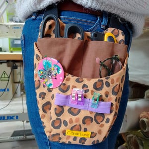 May include: A leopard print sewing tool organizer worn on the waist, featuring multiple pockets holding scissors, pins, and other craft supplies. The organizer has a purple elastic band with clips and a label that reads "Citron Lune."