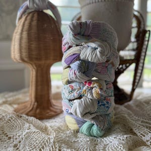 Op de afbeelding: Een stapel stoffen haarbanden in verschillende pastelkleuren, waaronder wit, lavendel en geel. Links staat een geweven rotan hoofdvorm. De haarbanden lijken gemaakt van hergebruikte stof.