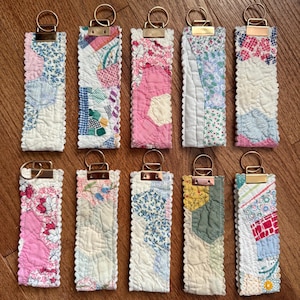 Könnte beinhalten: Zehn rechteckige Schlüsselanhänger aus Stoff mit verschiedenen Patchwork-Steppmuster in verschiedenen Farben wie Rosa, Blau, Grün und Weiß. Jeder Anhänger hat einen gewellten Rand und ist an einem goldfarbenen Metallring befestigt.
