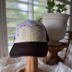Könnte beinhalten: Eine Trucker-Cap mit dunkelbraunem Schirm und weißem Vorderteil. Das Vorderteil ist mit einem Patchwork-Design verziert, das aus hellgelbem Stoff und floral gemusterten Stoffstücken besteht, die in einem geometrischen Muster vernäht sind.