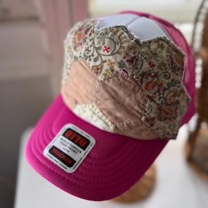 May include: A pink trucker hat with a floral patchwork design on the crown. The hat features a curved brim and a white mesh back. The label on the brim reads "OTTO WORLD FAMOUS FOAM FRONT TRUCKER".
