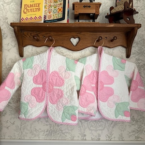 May include: Two matching quilted jackets featuring a white base with large pink flower petal appliques and light green leaf accents. The jackets have long sleeves, a rounded open front, and are finished with a pink and white checkered trim.