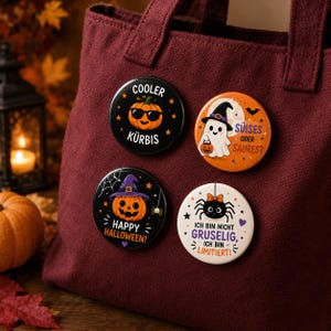 Könnte beinhalten: Vier runde Ansteckbuttons an einer weinroten Stofftasche. Die Buttons zeigen Halloween-Motive: einen Kürbis mit Sonnenbrille COOLER KÜRBIS, ein Gespenst mit Hexenhut SÜISES ODER SAURES, einen Kürbis HAPPY HALLOWEEN und eine Spinne mit dem Text ICH BIN NICHT GRUSELIG, ICH BIN LIMITIERT.
