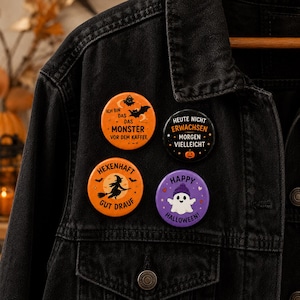 Könnte beinhalten: Vier runde Ansteckbuttons an einer schwarzen Jeansjacke. Die Buttons zeigen Halloween-Motive: ein orangefarbener mit Fledermaus und Text, ein schwarzer mit orangefarbenem Text und Kürbis, ein orangefarbener mit Hexe sowie ein lila Button mit einem Geist.