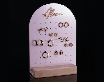 May include: Pink earring display stand with a wooden base. The stand has a cutout arch at the top with the word "Alma" written in gold lettering. The stand has multiple holes for displaying earrings.