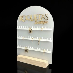 Puede incluir: Expositor de joyer&iacute;a de acr&iacute;lico blanco con el texto "KOQUETAS Boutique" en letras doradas. El expositor tiene m&uacute;ltiples filas de ganchos para colgar pendientes y otras joyas.