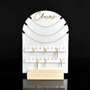 May include: White acrylic jewellery display stand with the word "Charms" in gold lettering. The stand has multiple rows of hooks for hanging necklaces and charms.